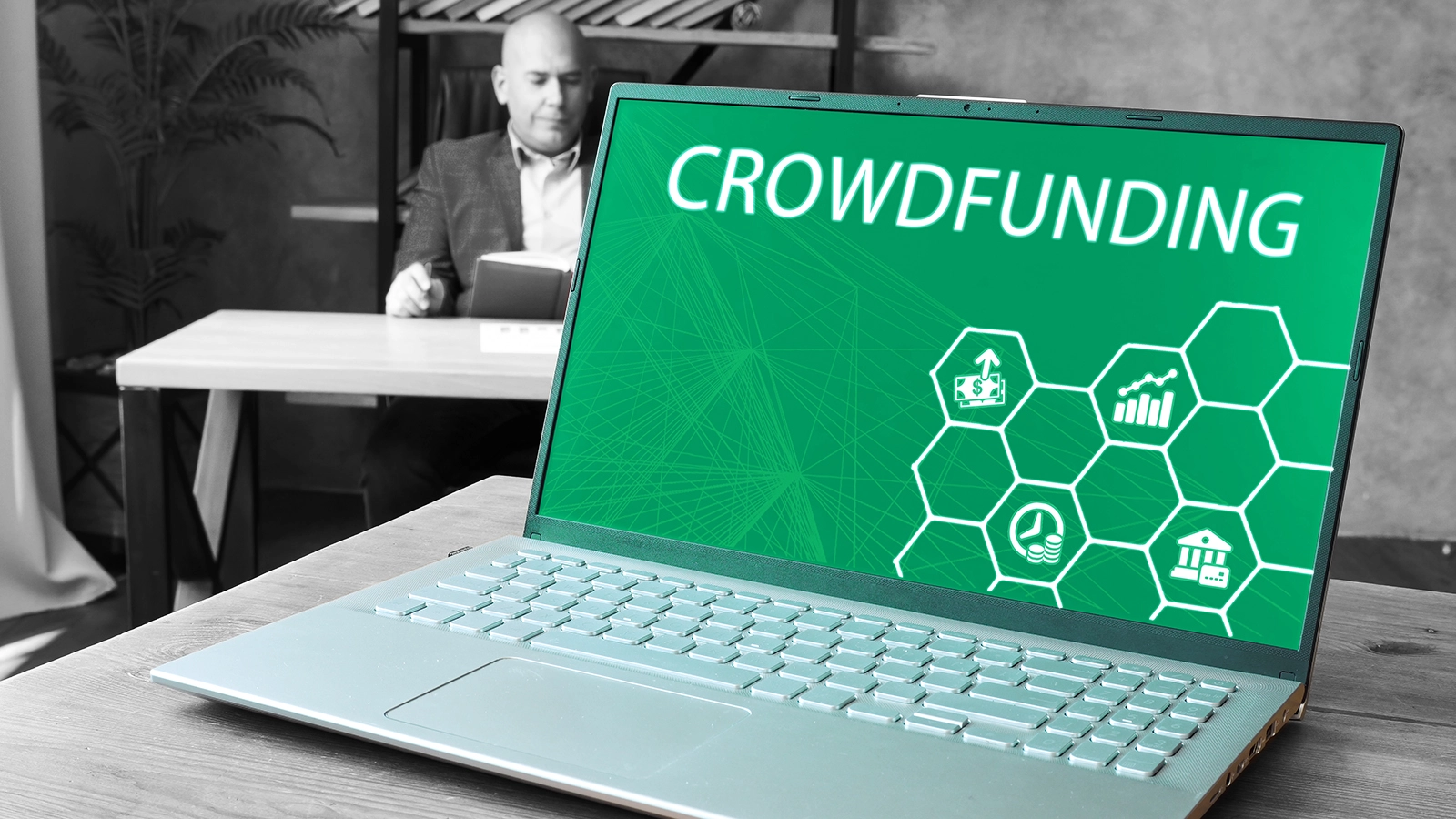 Ein Laptop steht auf dem Tisch, auf dem Display ist zu sehen Crowdfunding. Im Hintergrund sitzt ein Mann und ist entspannt.